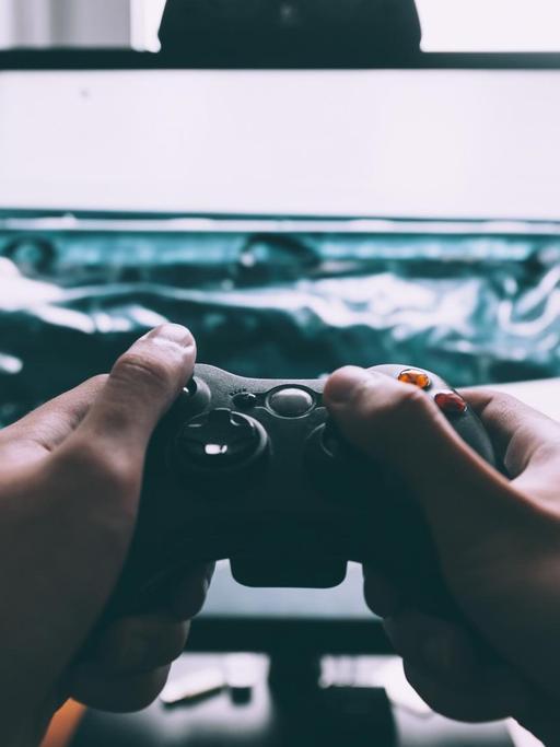 Ein Fernseher, auf dem ein Computerspiel läuft, im Vordergrund Hände, die eine Spielekonsole halten.