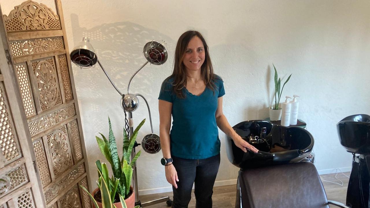 Eine junge Frau in T-Shirt und Jeans steht neben einem dunklen Friseursstuhl mit Waschbecken, auf das sie ihre Hand legt. Eine junge Frau in T-Shirt und Jeans steht neben einem dunklen Friseursstuhl mit Waschbecken, auf das sie ihre Hand legt.