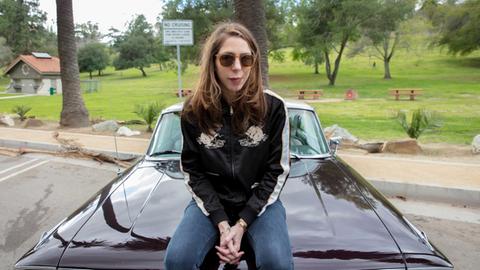 Rachel Kushner sitzt auf der Motorhaube eines us-amerikanischen Sportwagens. Sie trägt eine Sonnenbrille.
