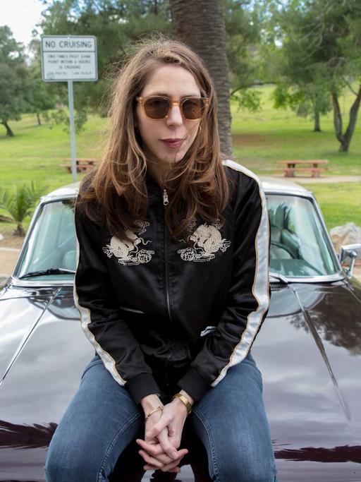 Rachel Kushner sitzt auf der Motorhaube eines us-amerikanischen Sportwagens. Sie trägt eine Sonnenbrille.
