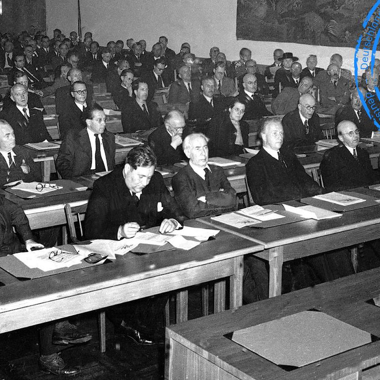 Erste Tagung des Parlamentarischen Rates am Nachmittag des 1. September 1948 in Bonn: Neben den 65 stimmberechtigten Mitgliedern aus westdeutschen Ländern nahmen Vertreter Berlins mit beratender Stimme an der Sitzung teil. In der ersten Reihe von links nach rechts: Max Reimann, KPD; Walter Menzel, SPD; Carlo Schmid, SPD; Theodor Heuss, FDP; Hans-Christoph Seebohm, DP; unbekannt; Adolf Suesterhenn, CDU; Anton Pfeiffer, CDU; Konrad Adenauer, CDU Erste Tagung des Parlamentarischen Rates am Nachmittag des 1. September 1948 in Bonn: Neben den 65 stimmberechtigten Mitgliedern aus westdeutschen Ländern nahmen Vertreter Berlins mit beratender Stimme an der Sitzung teil. In der ersten Reihe von links nach rechts: Max Reimann, KPD; Walter Menzel, SPD; Carlo Schmid, SPD; Theodor Heuss, FDP; Hans-Christoph Seebohm, DP; unbekannt; Adolf Suesterhenn, CDU; Anton Pfeiffer, CDU; Konrad Adenauer, CDU