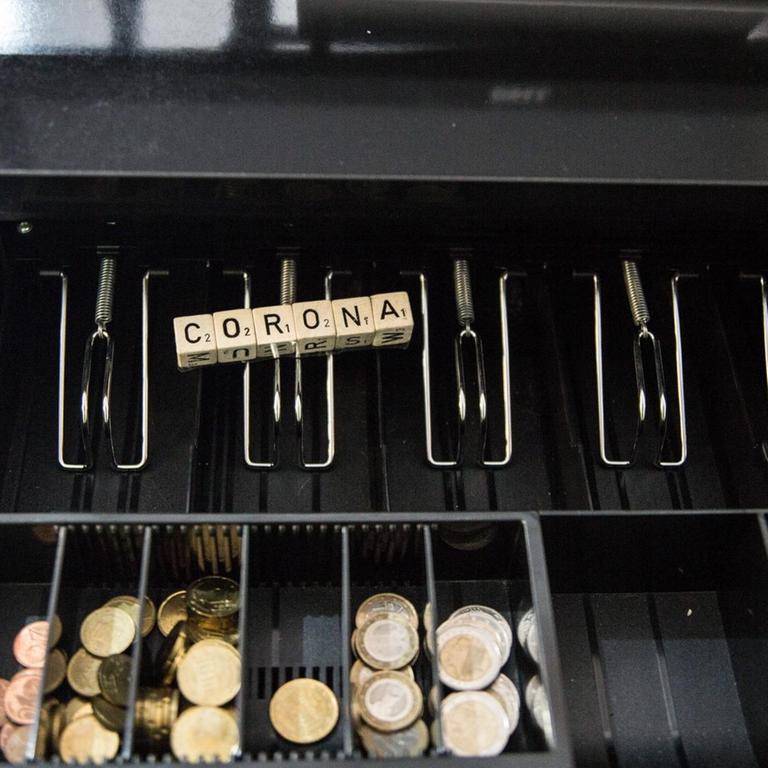 In der offenen Schublade einer Kasse liegt etwas Kleingeld, die Fächer für die Geldscheine sind leer. Auf den Fächern liegt der Schriftzug Corona.