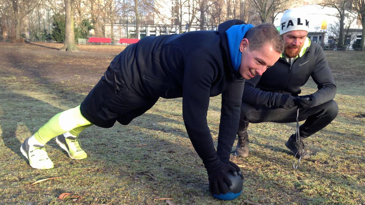 Ein Mann in Trainingskleidung absolviert auf einen Ball gestützt Fitnessübungen im Park. Neben ihm hockt sein Trainer Michel Gleich in Laufkleidung und mit einer Stoppuhr. Ein Mann in Trainingskleidung absolviert auf einen Ball gestützt Fitnessübungen im Park. Neben ihm hockt sein Trainer Michel Gleich in Laufkleidung und mit einer Stoppuhr.