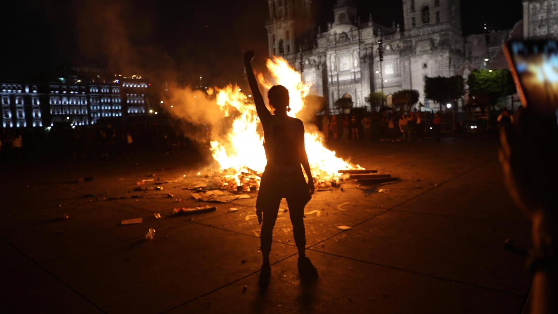 Silhouette eine Frau, die mit erhobenem rechten Arm im Dunkeln vor einem Feuer steht, das auf einem Platz in Mexiko City brennt.