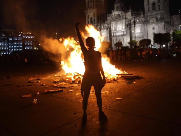 Silhouette eine Frau, die mit erhobenem rechten Arm im Dunkeln vor einem Feuer steht, das auf einem Platz in Mexiko City brennt.