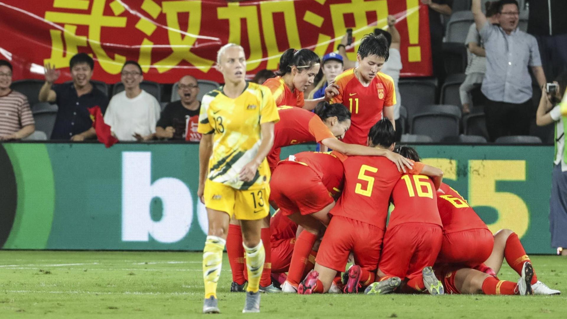 Die chinesische Fußballolympiamannschaft feiert ein Tor gegen Australien. Im Hintergrund chinesische Fans mit einem Transparent in Rot mit gelber Schrift.