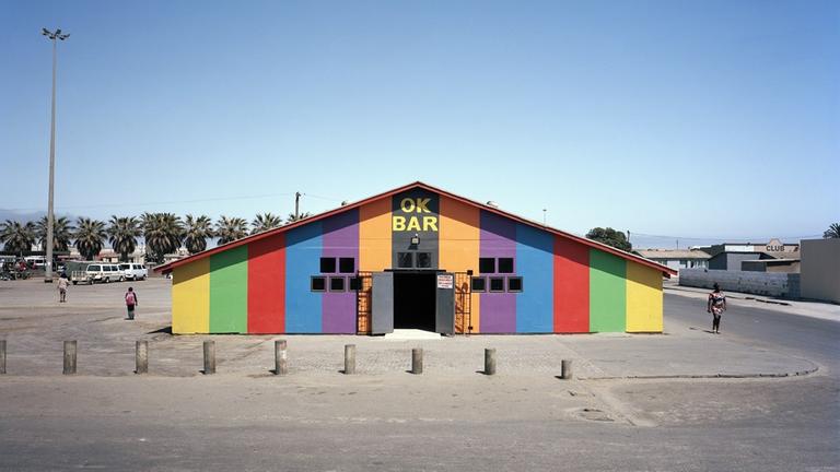 Eine Halle mit bunten Längsstreifen steht mitten auf einem großen Platz, ganz oben steht in gelben fetten Lettern 'OK BAR'. Eine Halle mit bunten Längsstreifen steht mitten auf einem großen Platz, ganz oben steht in gelben fetten Lettern 'OK BAR'.