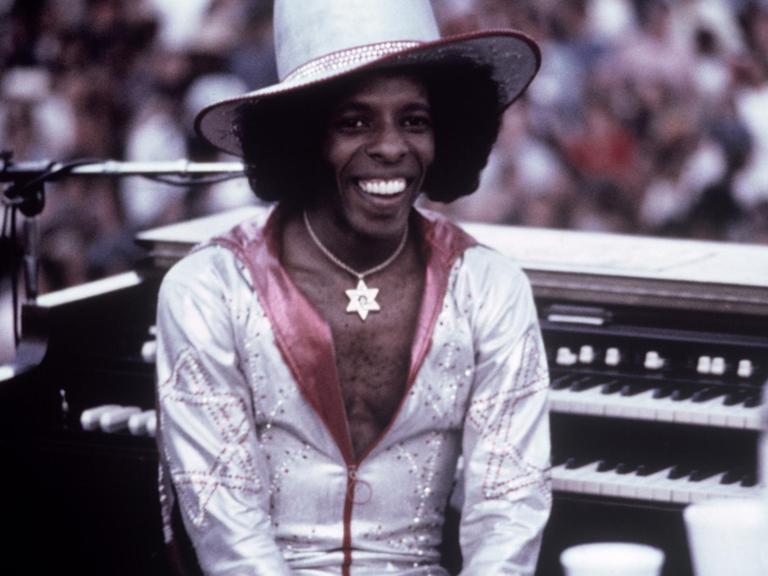 Der US-Musiker Sly Stone sitzt 1974 in einem weißglänzenden Anzug bei einem Auftritt auf einer Bühne.
