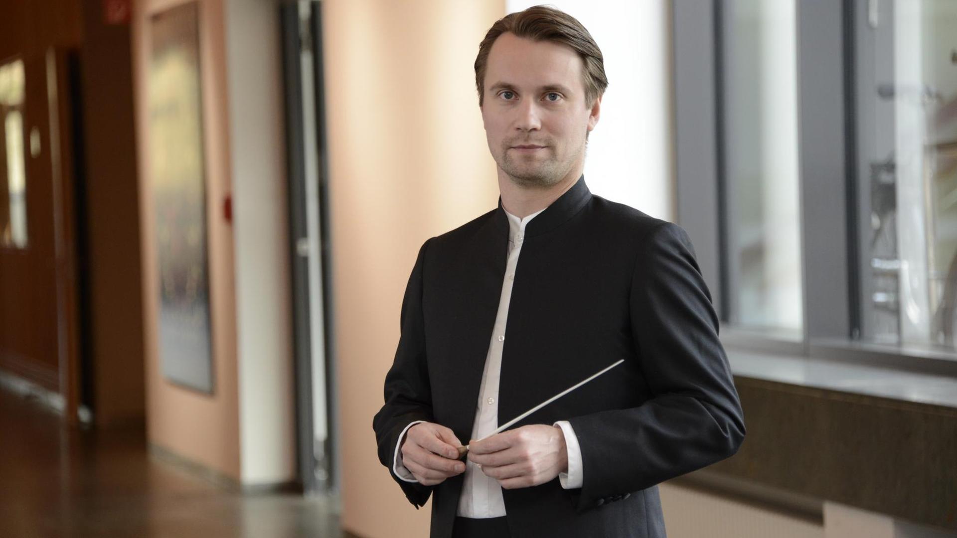 Ein Mann steht aufrecht in einem weitläufigen Flur, schaut in die Kamera und hält seinen Taktstock vor sich in seinen Händen. Ein Mann steht aufrecht in einem weitläufigen Flur, schaut in die Kamera und hält seinen Taktstock vor sich in seinen Händen.