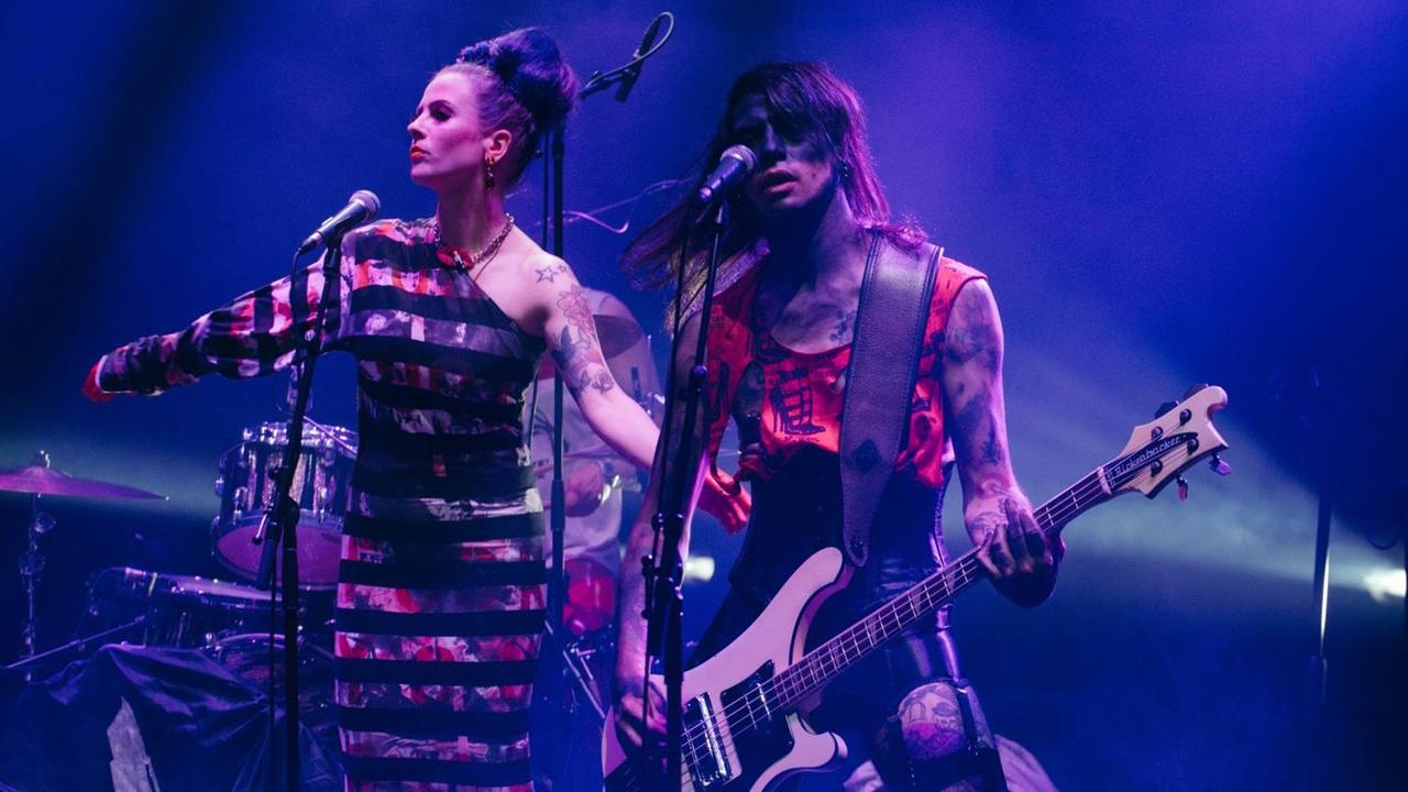 Der geschminkte und stark tätowierte Bassist von "Eat Lipstick" performt neben der Sängerin auf einer blau angestrahlten Bühne. Der geschminkte und stark tätowierte Bassist von "Eat Lipstick" performt neben der Sängerin auf einer blau angestrahlten Bühne.
