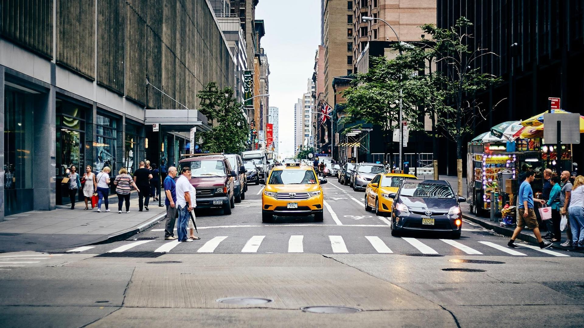 Blick auf einen Fußübergang in New York. Am Rand stehen Menschen. Auf der Straße eines der gelben Taxi