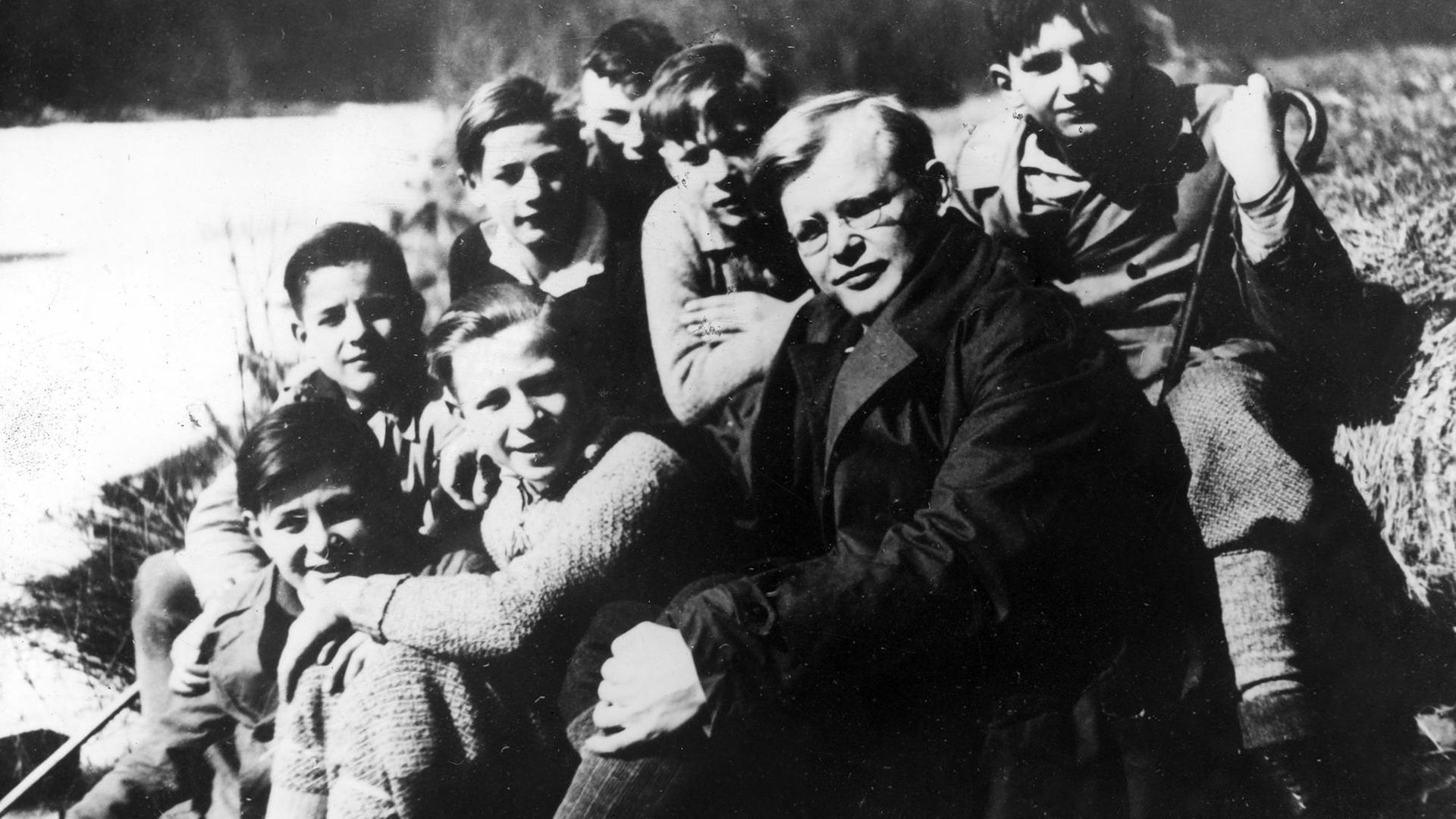 Dietrich Bonhoeffer sitzt inmitten einer Gruppe Jungen an einem Flussufer, alle schauen in die Kamera.
