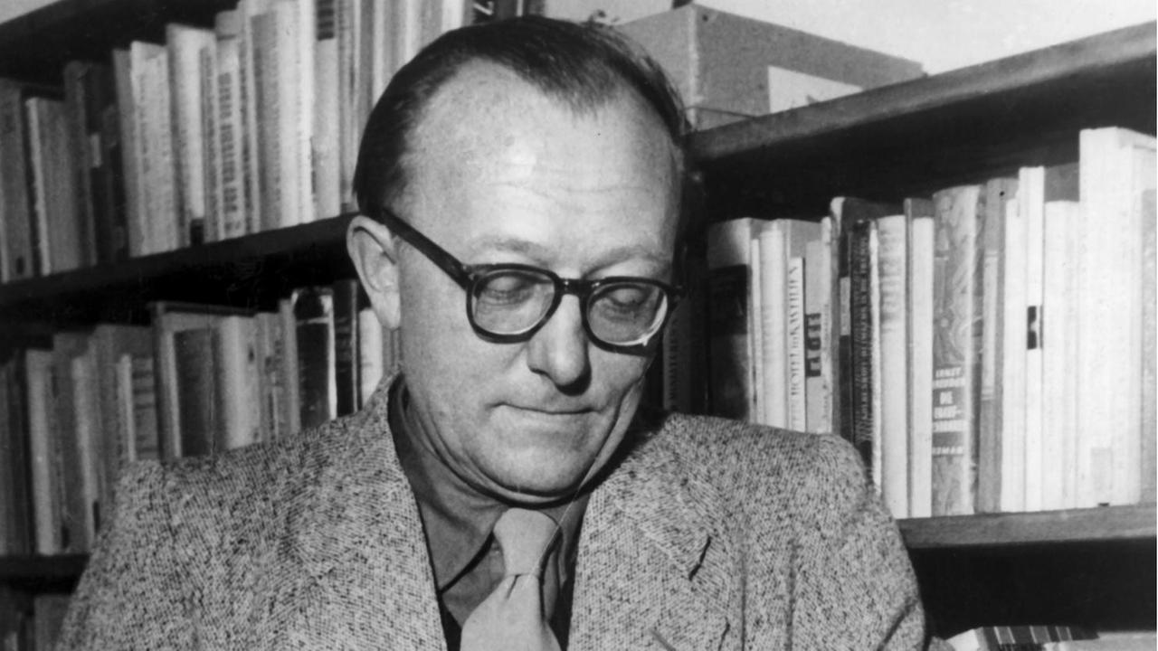 Der deutsche Schriftsteller Günter Eich lehnt an einem Regal und blättert in einem Buch. Aufnahme von 1953.