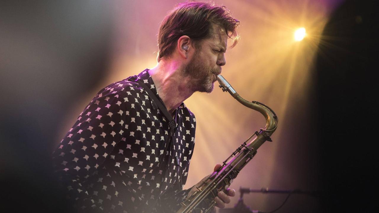 Im Gegenlicht eines Bühnenscheinwerfers spielt ein bärtiger Mann mittleren Alters Tenorsaxofon. Im Gegenlicht eines Bühnenscheinwerfers spielt ein bärtiger Mann mittleren Alters Tenorsaxofon.