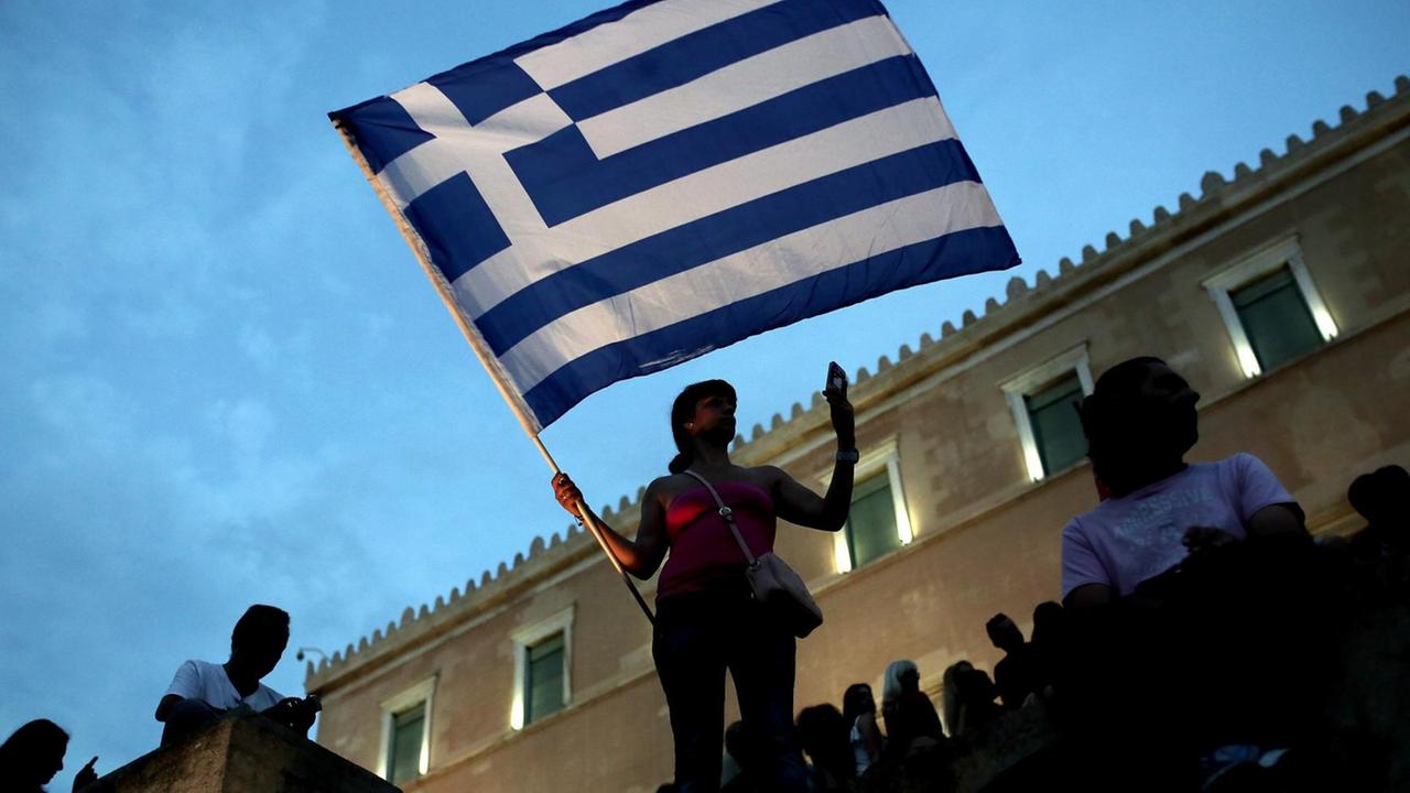 Eine Frau steht während der Demonstration auf einer Mauer vor dem Abendhimmel. Sie hält eine griechische Flagge. Eine Frau steht während der Demonstration auf einer Mauer vor dem Abendhimmel. Sie hält eine griechische Flagge.