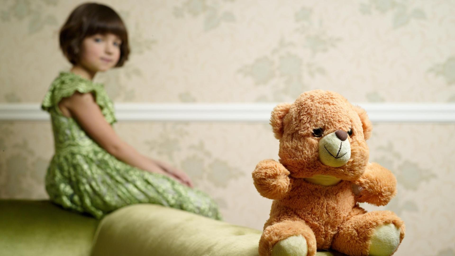 Ein alter Teddy sitzt auf einer Sofalehne, im Hintergrund ist ein Mädchen zu sehen. Ein alter Teddy sitzt auf einer Sofalehne, im Hintergrund ist ein Mädchen zu sehen.