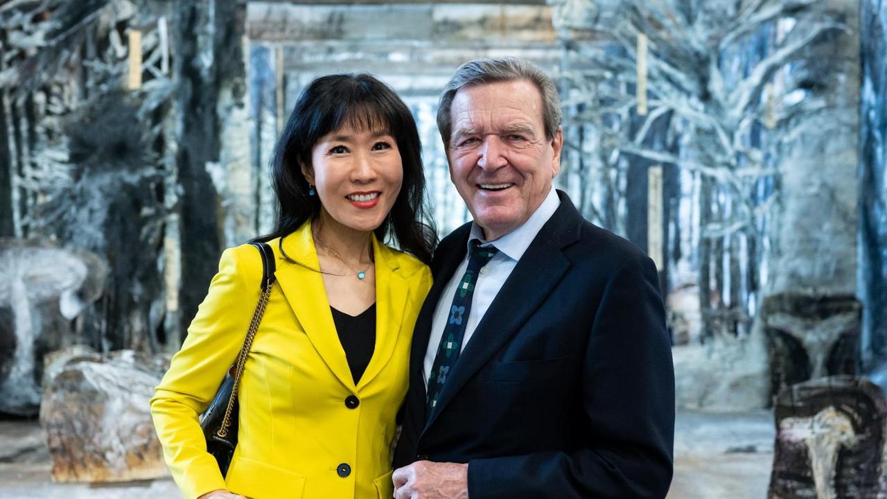 Eine Frau in knallgelbem Hosenanzug und ein Mann in dunkelblauem Anzug halten sich im Arm und posieren vor einem Bühnenbild für den Fotografen. Es handelt sich um Alt-Bundeskanzler Gerhard Schröder und seine Frau Soyeon Schröder-Kim, sie stehen vor einem Bühnenbild-Projekt von Anselm Kiefer. Eine Frau in knallgelbem Hosenanzug und ein Mann in dunkelblauem Anzug halten sich im Arm und posieren vor einem Bühnenbild für den Fotografen. Es handelt sich um Alt-Bundeskanzler Gerhard Schröder und seine Frau Soyeon Schröder-Kim, sie stehen vor einem Bühnenbild-Projekt von Anselm Kiefer.