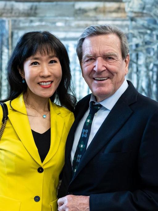 Eine Frau in knallgelbem Hosenanzug und ein Mann in dunkelblauem Anzug halten sich im Arm und posieren vor einem Bühnenbild für den Fotografen. Es handelt sich um Alt-Bundeskanzler Gerhard Schröder und seine Frau Soyeon Schröder-Kim, sie stehen vor einem Bühnenbild-Projekt von Anselm Kiefer.