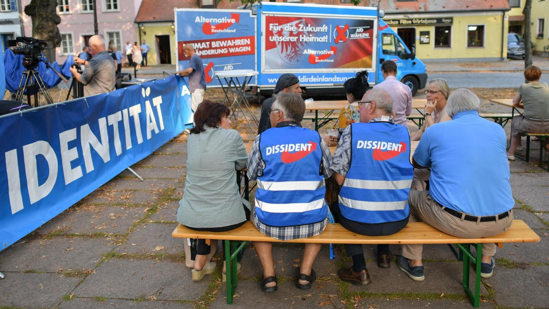 Mehrere Senioren sitzen in AfD-Westen mit dem Aufdruck "Dissident" auf einer Bank mit dem Rücken zum Betrachter. Auf einem Banner an der linken Seite steht "Identität". Mehrere Senioren sitzen in AfD-Westen mit dem Aufdruck "Dissident" auf einer Bank mit dem Rücken zum Betrachter. Auf einem Banner an der linken Seite steht "Identität".