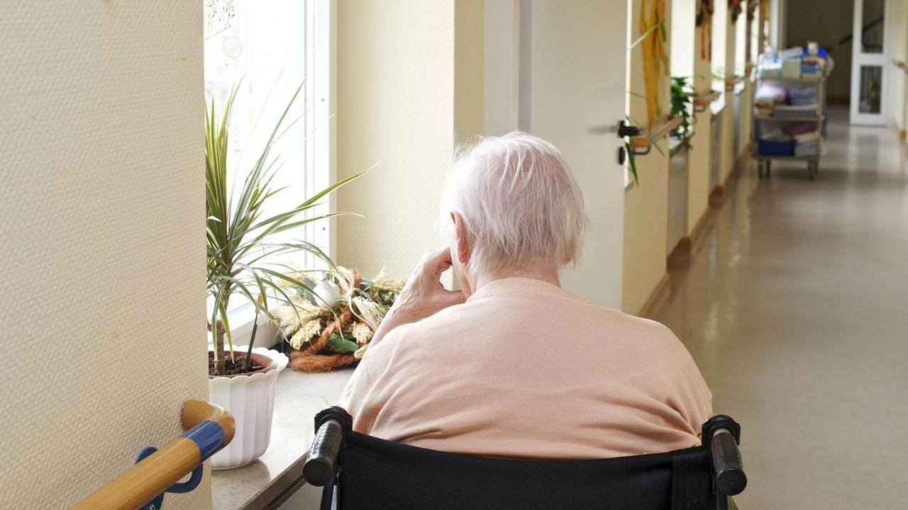 Eine Seniorin im Rollstuhl sitzt alleine im leeren Flur eines Altenheimes und schaut aus dem Fenster.