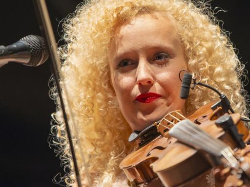 Eine Frau mit blonden gelockten Harren steht auf einer Bühne und spielt Geige.
