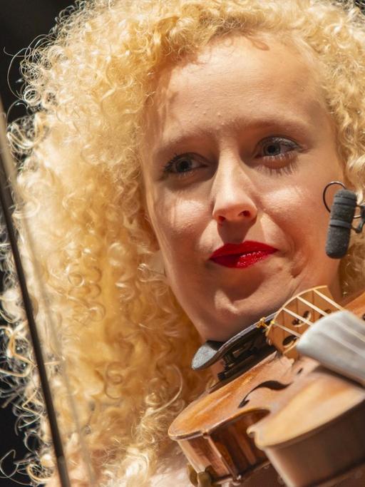 Eine Frau mit blonden gelockten Harren steht auf einer Bühne und spielt Geige.