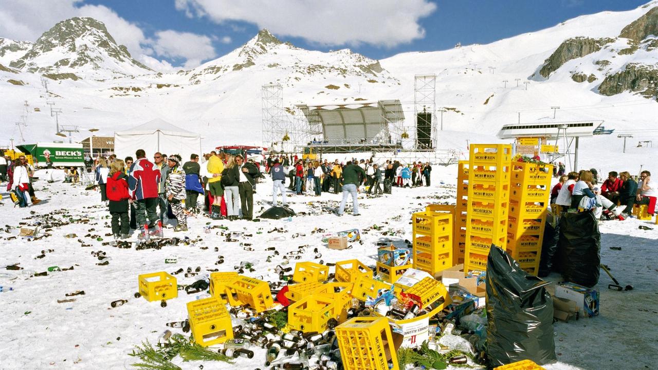 Feiernde Menschen in Ischgl, inmitten der Schneelandschaft liegen die Reste der Feier und gelbe Kartons.