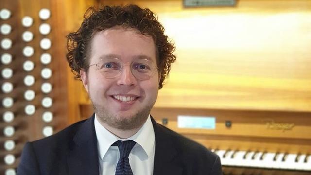 Ein Mann mit runder Brille, leichtem Vollbart und lockigem Haar sitzt vor dem Spieltisch einer Orgel.