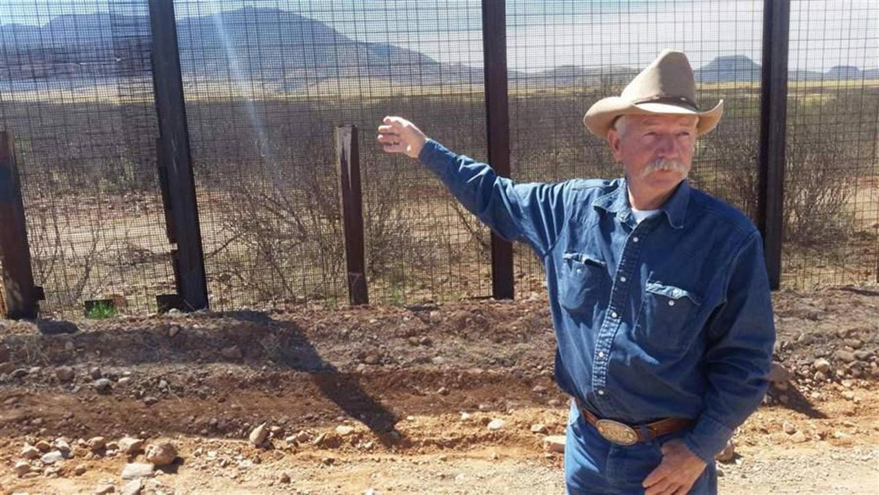 John Ladd mit Schnäuzer und Cowboyhut zeigt auf einen Grenzzaun, im Hintergrund Berge John Ladd mit Schnäuzer und Cowboyhut zeigt auf einen Grenzzaun, im Hintergrund Berge