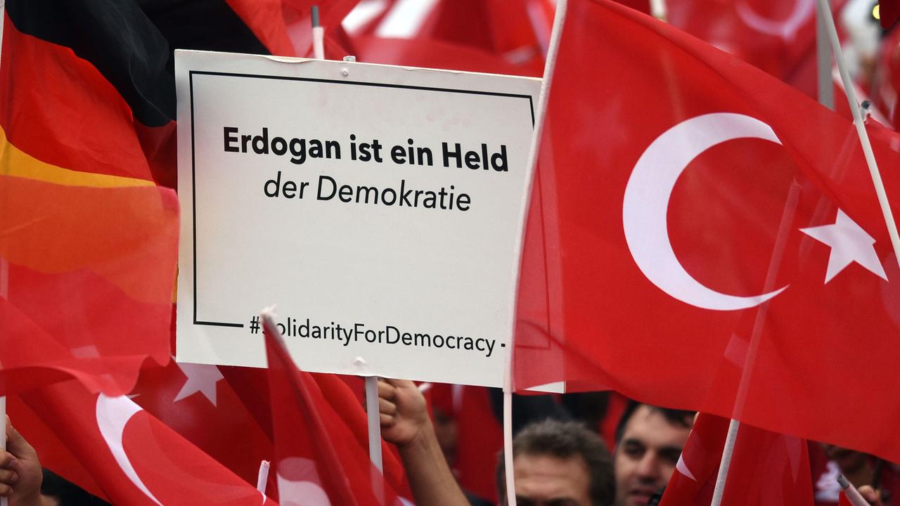 Anhänger des türkischen Staatspräsidenten Erdogan halten in Köln Fahnen. Einer hält ein Schild hoch mit der Aufschrift "Erdogan ist ein Held der Demokratie".
