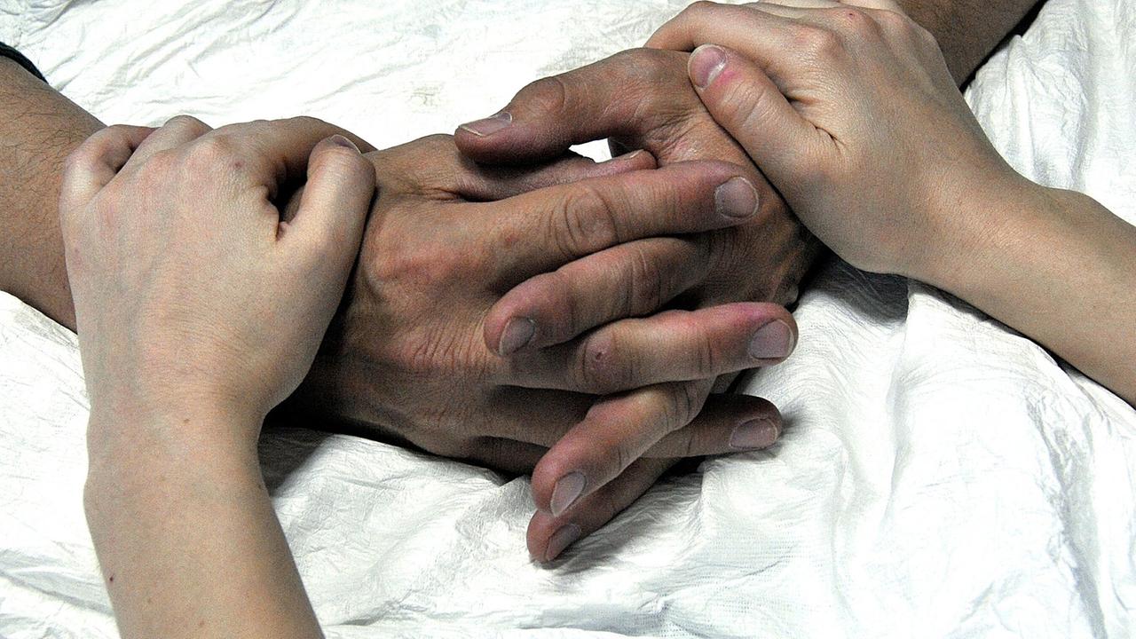Die Hände eines alten Menschen im Krankenhausbett werden von einer jüngeren Person gehalten. Die Hände eines alten Menschen im Krankenhausbett werden von einer jüngeren Person gehalten.