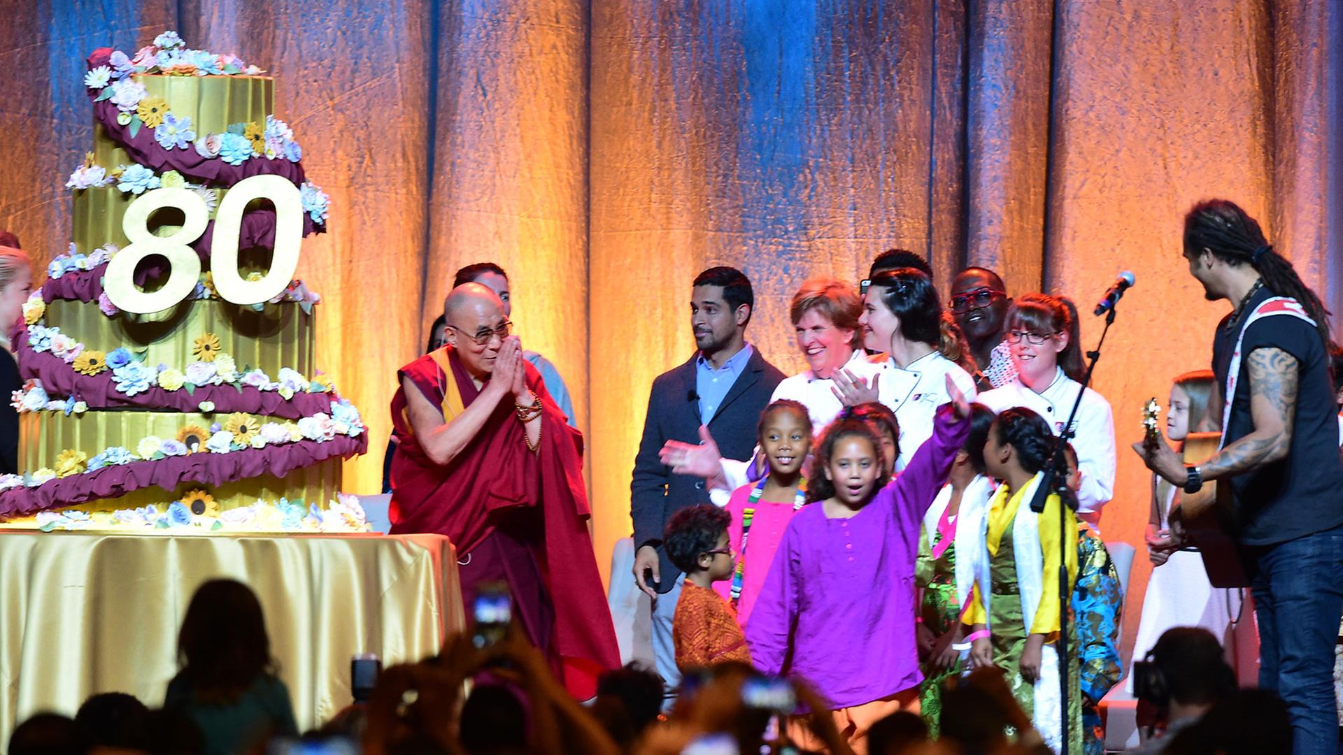 Der Dalai Lama steht auf einer Bühne, neben ihm eine übermannshohe Geburtstagstorte mit einer großen 80., der Musiker Michael Frente Kinder. Der Dalai Lama steht auf einer Bühne, neben ihm eine übermannshohe Geburtstagstorte mit einer großen 80., der Musiker Michael Frente Kinder.