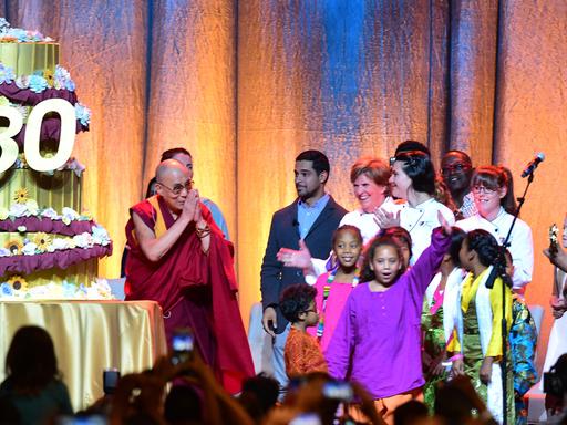 Der Dalai Lama steht auf einer Bühne, neben ihm eine übermannshohe Geburtstagstorte mit einer großen 80., der Musiker Michael Frente Kinder.