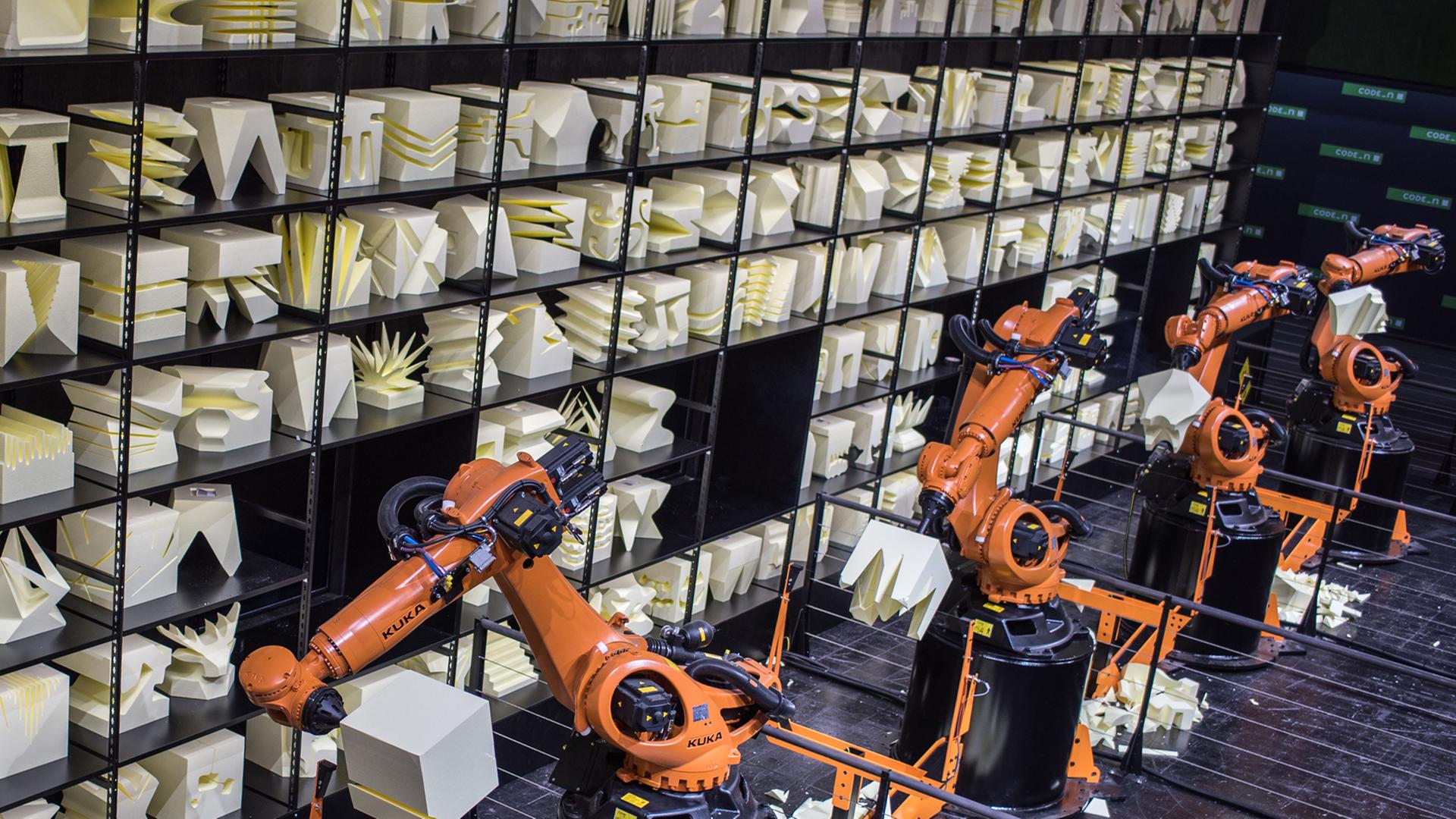 Orangefarbener Produktionsroboterarm, im Hintergrund ein Regal mit Kunstobjekten aus Holz. Orangefarbener Produktionsroboterarm, im Hintergrund ein Regal mit Kunstobjekten aus Holz.