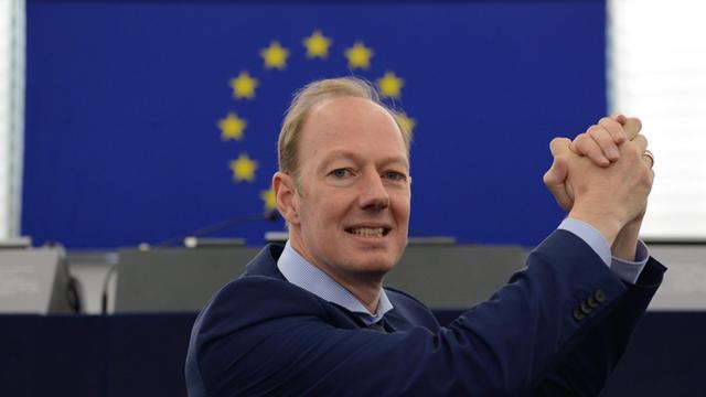 Journalist, Satiriker und Mitglied des Europäischen Parlaments, Martin Sonneborn, macht eine Siegerpose mit den Händen, dabei leuchten die Sterne der EU-Flagge über seinem Kopf