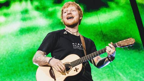 Ed Sheeran steht mit einer Gitarre auf der Bühne, der Hintergrund ist grün.