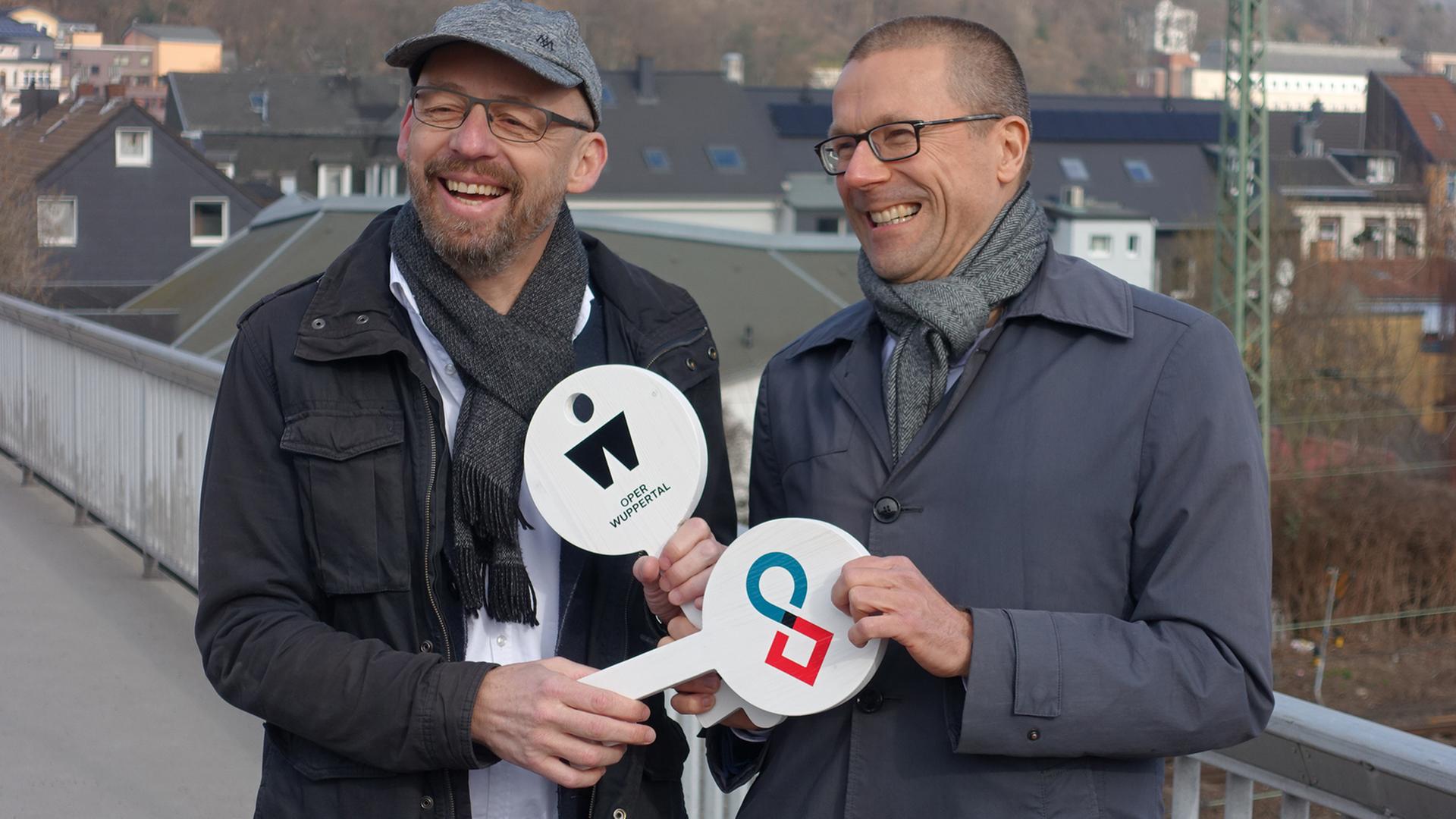 Berthold Schneider (links) und Uwe Schneidewind halten symbolische weiße Schlüssel aus Holz in den Händen, auf denen das Symbol der Oper Wuppertal und des Wuppertal Instituts zu sehen sind.