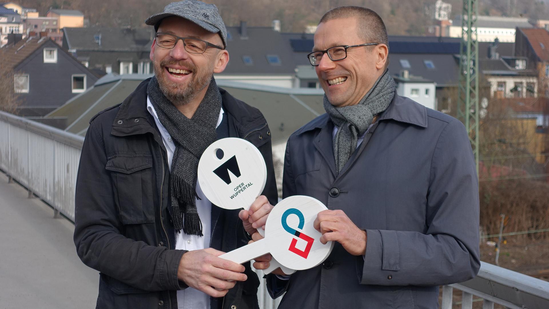 Berthold Schneider (links) und Uwe Schneidewind halten symbolische weiße Schlüssel aus Holz in den Händen, auf denen das Symbol der Oper Wuppertal und des Wuppertal Instituts zu sehen sind. Berthold Schneider (links) und Uwe Schneidewind halten symbolische weiße Schlüssel aus Holz in den Händen, auf denen das Symbol der Oper Wuppertal und des Wuppertal Instituts zu sehen sind.