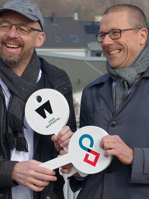 Berthold Schneider (links) und Uwe Schneidewind halten symbolische weiße Schlüssel aus Holz in den Händen, auf denen das Symbol der Oper Wuppertal und des Wuppertal Instituts zu sehen sind.