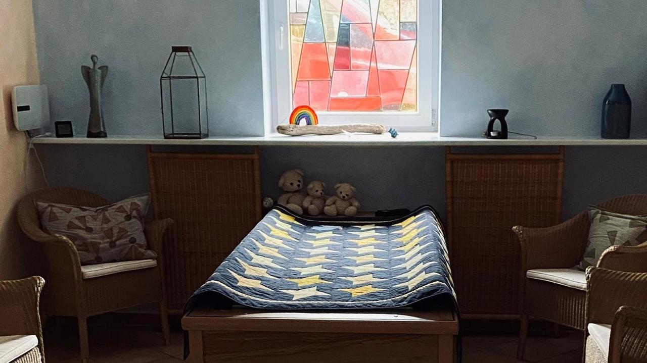 Vor einem bunten Glasfenster steht ein Bett mit einer Bahre darauf, auf der eine blau gelbe Decke liegt, daneben stehen vier Rattansessel.