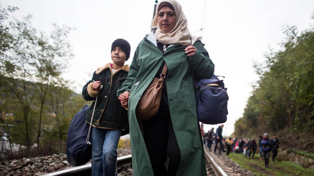 Die Frau mit Kopftuch hat einen Jungen an der Hand. Beide tragen Taschen. Im Hintergrund sind weitere Flüchtlinge an den Gleisen zu sehen.