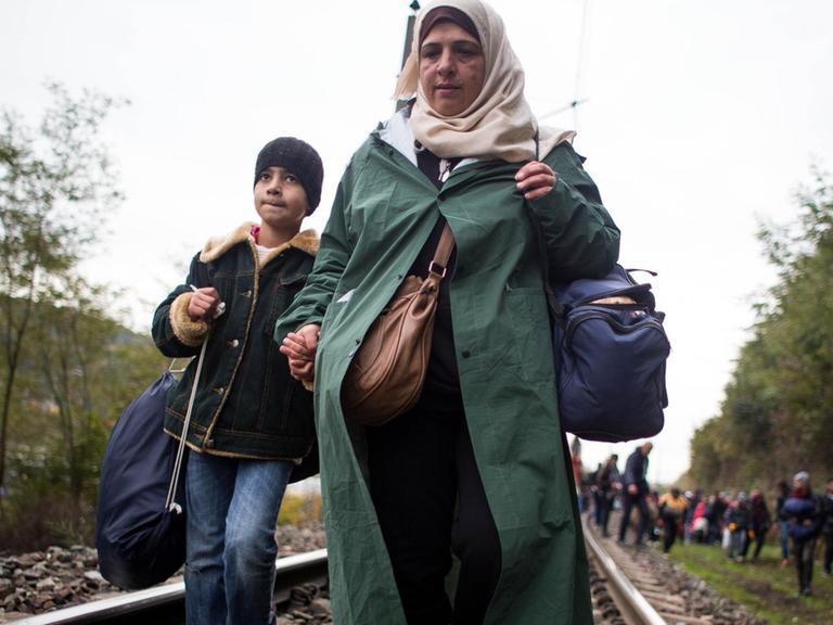Die Frau mit Kopftuch hat einen Jungen an der Hand. Beide tragen Taschen. Im Hintergrund sind weitere Flüchtlinge an den Gleisen zu sehen.