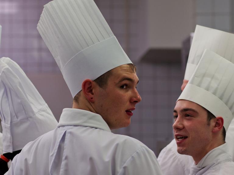 Mehrere Kochlehrlinge mit hohen weißen Kochmützen stehen bei einem Wettbewerb in einer Großküche.