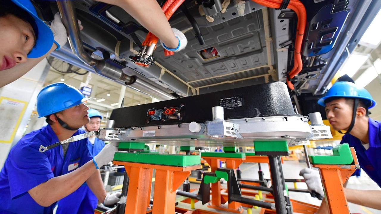 Unteransicht eines Elektroautos, in das eine Batterie installiert wird. Einige chinesische Arbeiter in blauen Fabrikmonturen stehen darum und legen Hand an oder beobachten das Geschehen interessiert.