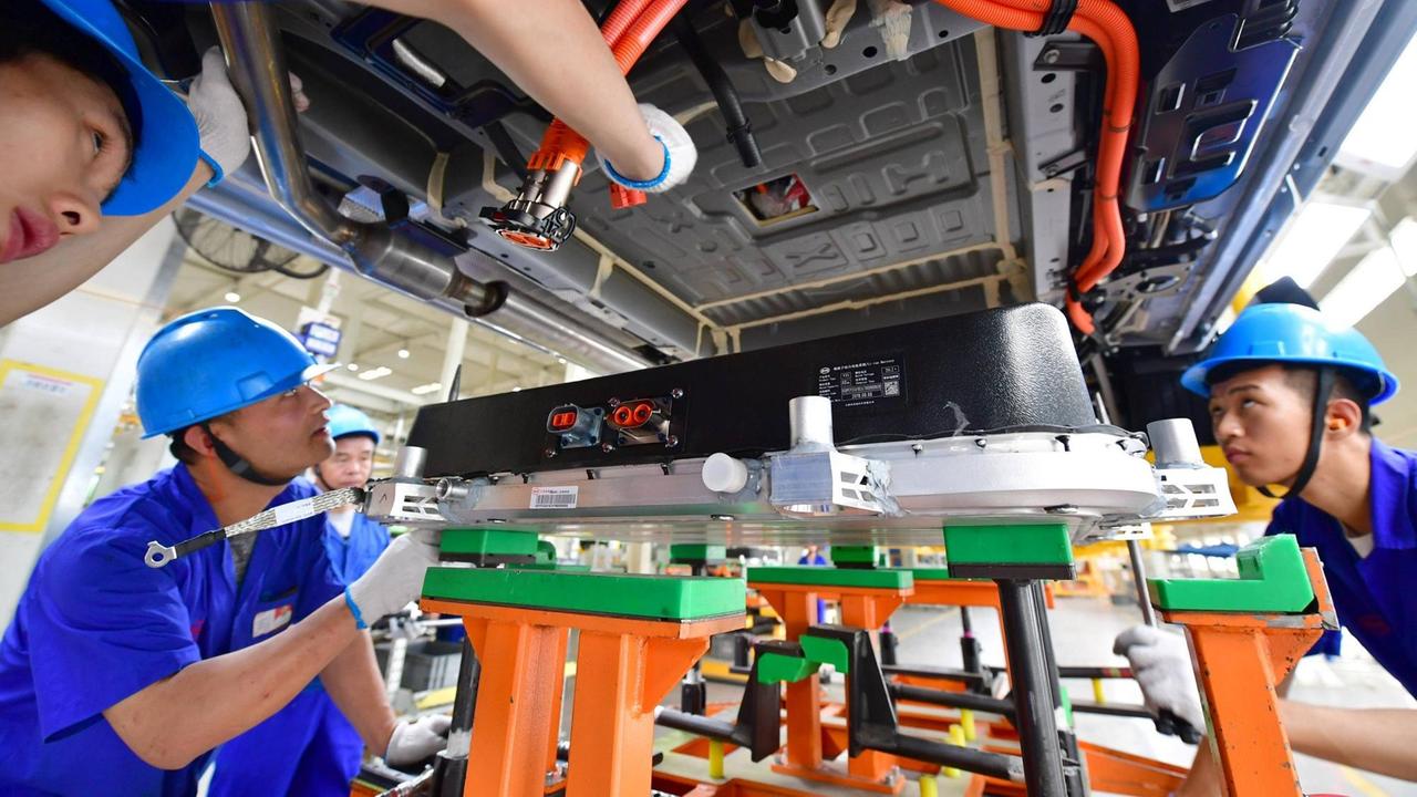 Unteransicht eines Elektroautos, in das eine Batterie installiert wird. Einige chinesische Arbeiter in blauen Fabrikmonturen stehen darum und legen Hand an oder beobachten das Geschehen interessiert. Unteransicht eines Elektroautos, in das eine Batterie installiert wird. Einige chinesische Arbeiter in blauen Fabrikmonturen stehen darum und legen Hand an oder beobachten das Geschehen interessiert.