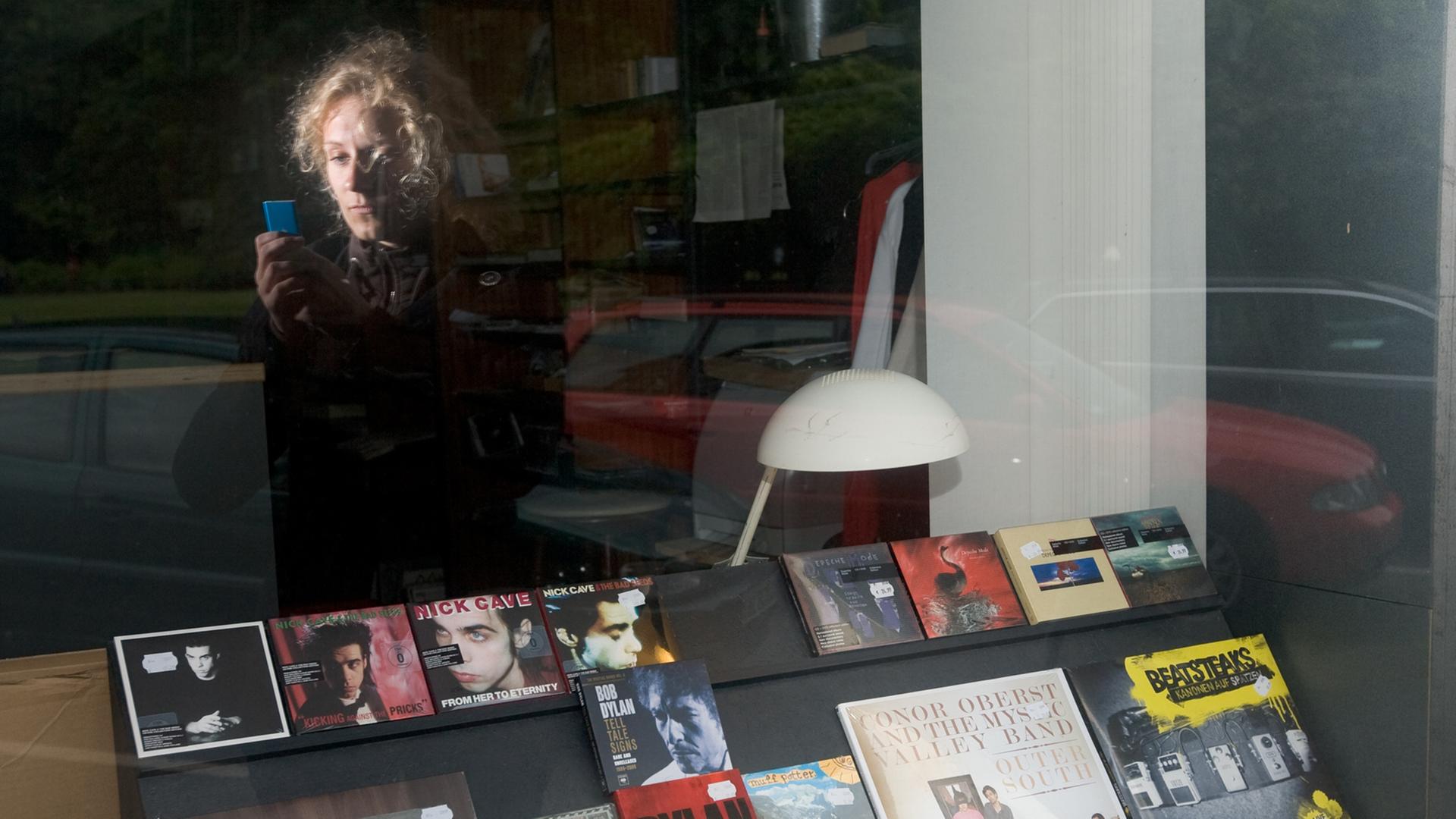 Eine junge Frau blickt auf ihren MP3-Player, während sie vor dem Schaufenster eines Plattenladens steht. Eine junge Frau blickt auf ihren MP3-Player, während sie vor dem Schaufenster eines Plattenladens steht.