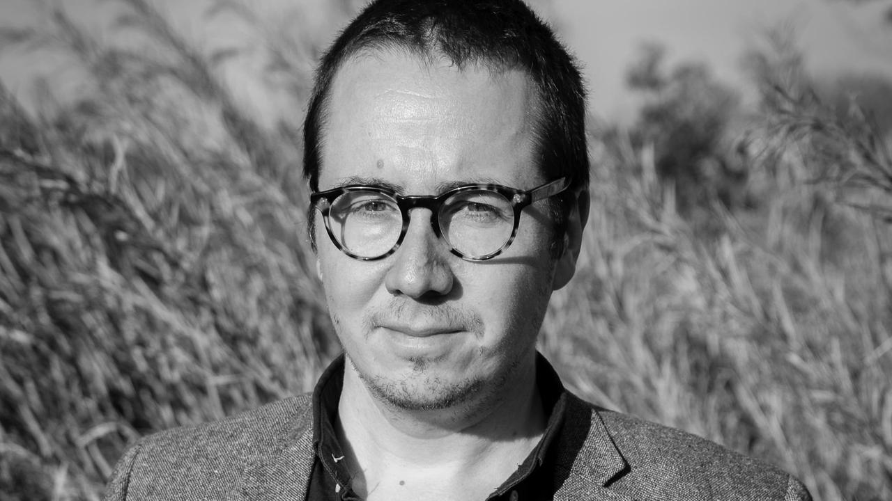 Schwarzweißaufnahme. Gabriel Wollner steht in einem Feld, trägt eine schwarze brille und ein Sakko. Schwarzweißaufnahme. Gabriel Wollner steht in einem Feld, trägt eine schwarze brille und ein Sakko.