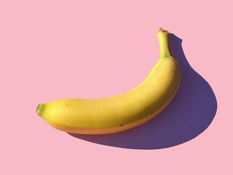 Eine Banane liegt auf einem rosa Untergrund und wirft einen scharfen Schatten darauf.