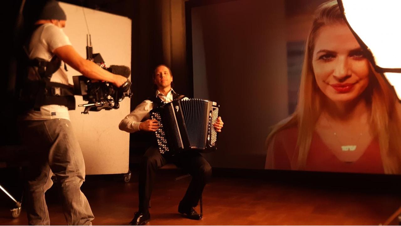 In der Mitte des Bildes sitzt der Akkordeonist Maxim Shagaev, der während er spielt in Richtung eines Kameramanns rechts von ihm blickt. Im Hintergrund hängt ein riesiges Portrait einer jungen Schauspielerin. In der Mitte des Bildes sitzt der Akkordeonist Maxim Shagaev, der während er spielt in Richtung eines Kameramanns rechts von ihm blickt. Im Hintergrund hängt ein riesiges Portrait einer jungen Schauspielerin.