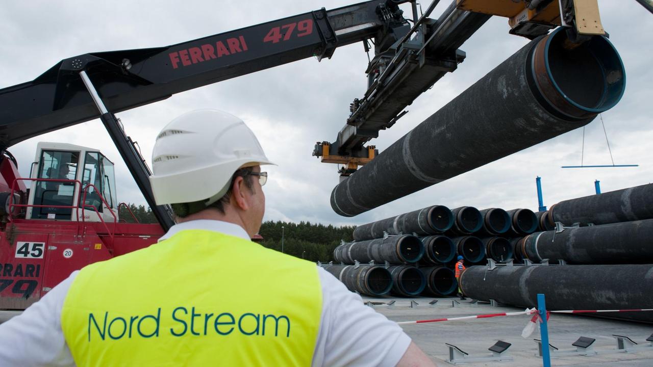 Im Vordergrund steht ein Mann in gelber Warnweste und beobachtet einen Bagger, der Pipelinerohre hebt. Im Vordergrund steht ein Mann in gelber Warnweste und beobachtet einen Bagger, der Pipelinerohre hebt.